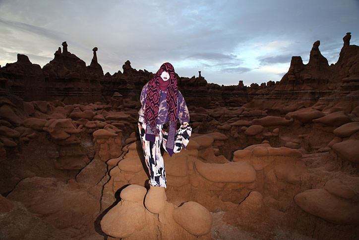 A person stands amidst a landscape of rounded, reddish-brown rock formations under a pale sky. They wear a purple patterned top, a long red and purple scarf, and wide-legged black and white trousers.