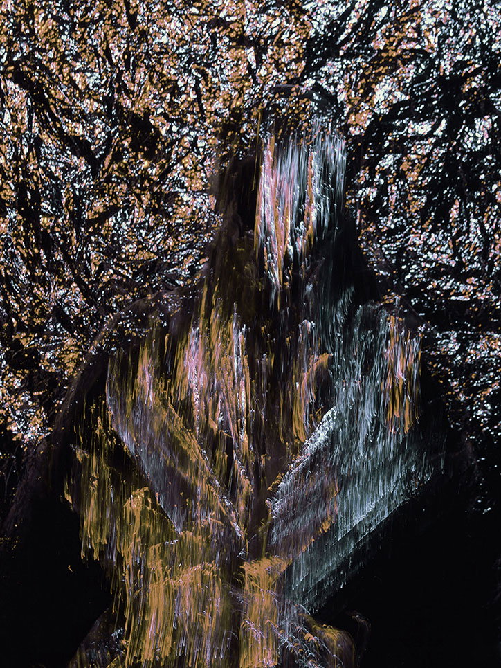 An abstract image of a blurred, shimmering figure composed of vertical streaks of gold, silver and purple light. The figure is set against a dark, textured background that resembles crushed foil or rocky terrain.