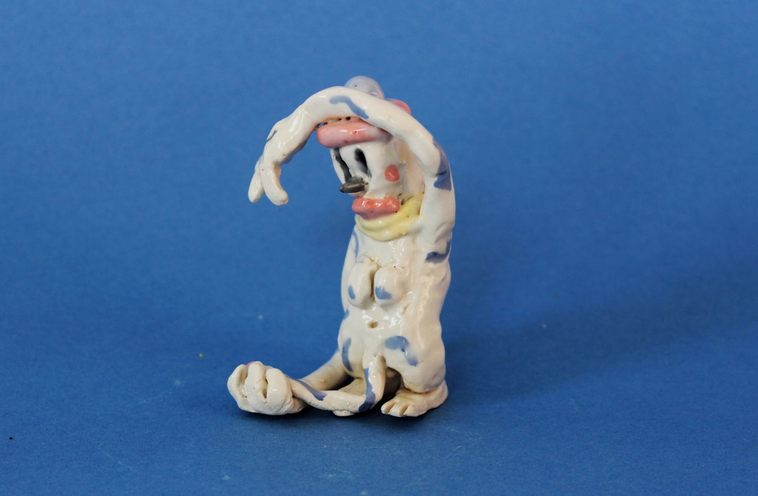 A small ceramic sculpture of a distorted, white figure with blue mottled patches. The figure is crouched with one arm raised over its head against a plain blue background.