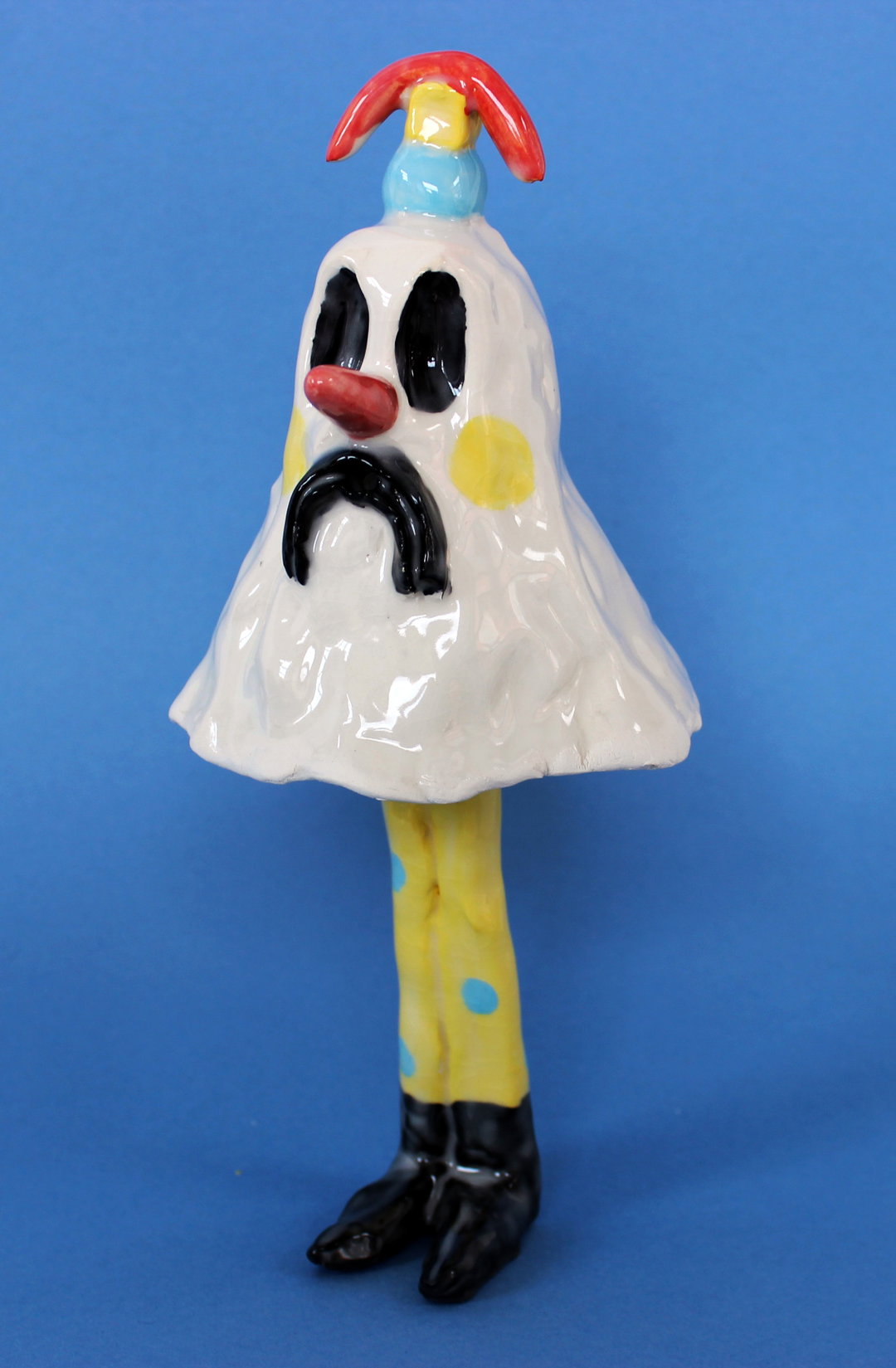 A ceramic sculpture of a sad clown against a plain blue background. The figure wears a white, bell-shaped tunic with a yellow polka dot and a red nose, topped with a small red and blue hat.
