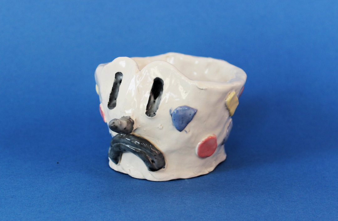 A small, white ceramic vessel with a glossy glaze and a distorted, face-like appearance. It features two narrow black slits for eyes, a small protruding nose, and a downturned black mouth.