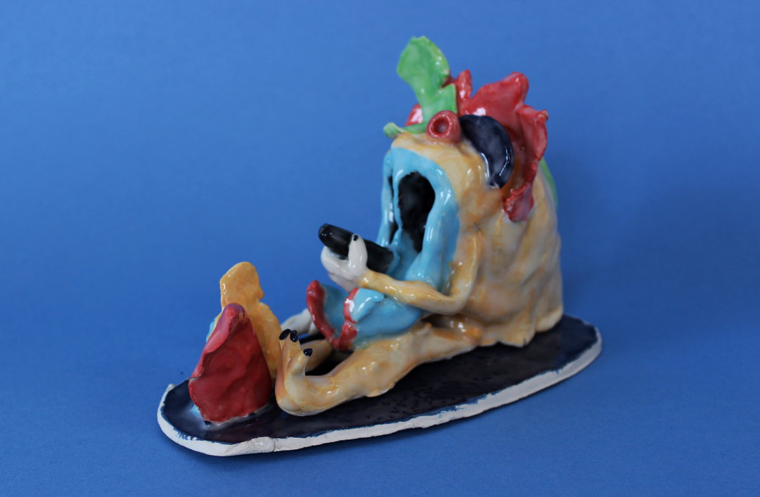 A ceramic sculpture of a slumped, melting figure in blue and yellow, holding a small black device. The figure is topped with red and green organic shapes and sits on a dark oval base against a blue background.