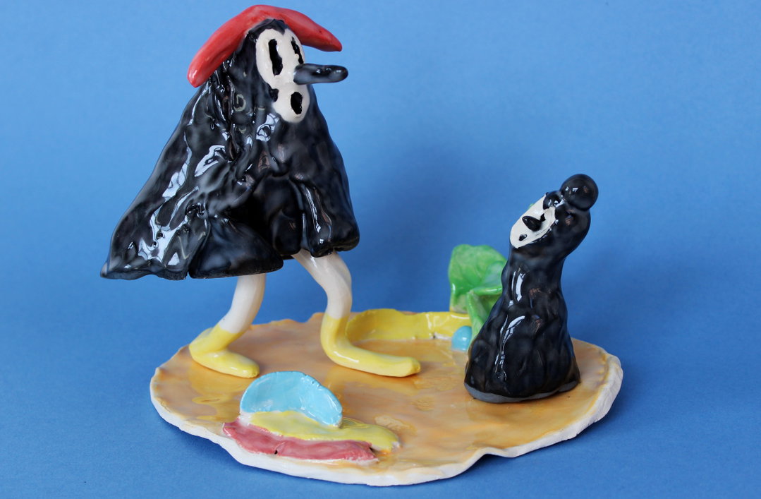 A ceramic sculpture of two glossy black figures with white faces on a yellow base. One figure wears a red hat and looks down at a smaller figure looking up.