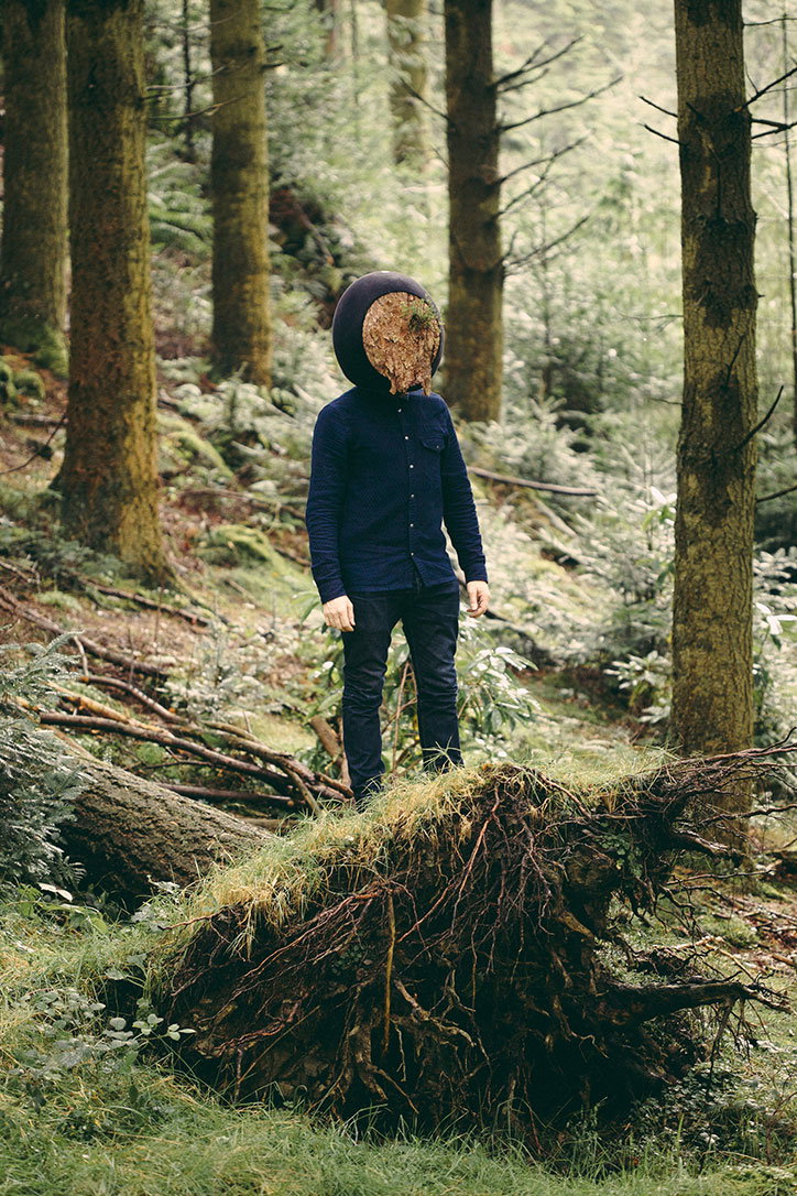 A person stands in a dense forest, wearing a dark blue shirt and trousers. Their head is replaced by a large, circular mass of dried grass and roots.