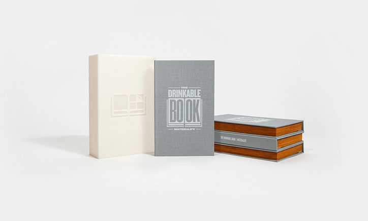 A white slipcase and a grey book stand beside a stack of three identical books. The cover of the grey book features the title “The Drinkable Book” and the phrase “Water is Life”.