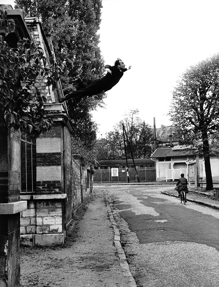 A person in dark clothing leaps horizontally from the roof of a stone building. Below them, a person on a bicycle rides down a quiet street.