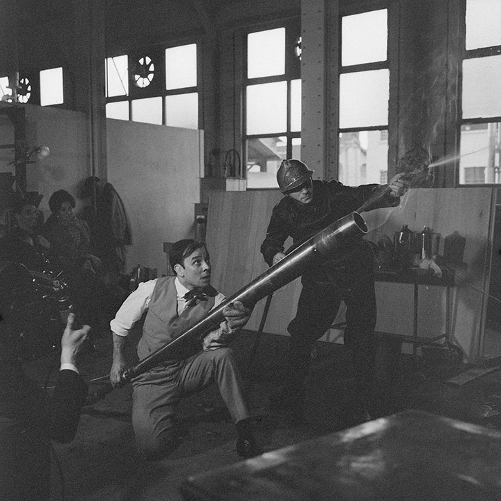 A black and white photo of two people in a workshop. One person crouches while holding a long metal tube, looking up in surprise at another person who is using a blowtorch.