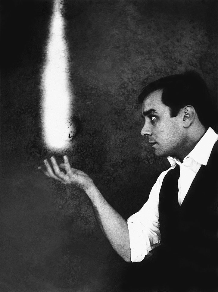 A person in a white shirt and dark waistcoat looks towards a bright, vertical beam of light. They hold their hand open beneath the glow against a dark, textured background.