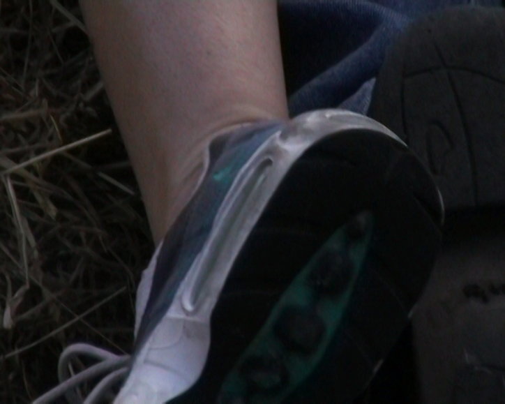 A close-up of a person's foot wearing a dark trainer with a white sole. They are resting against a background of dry grass and dark fabric.