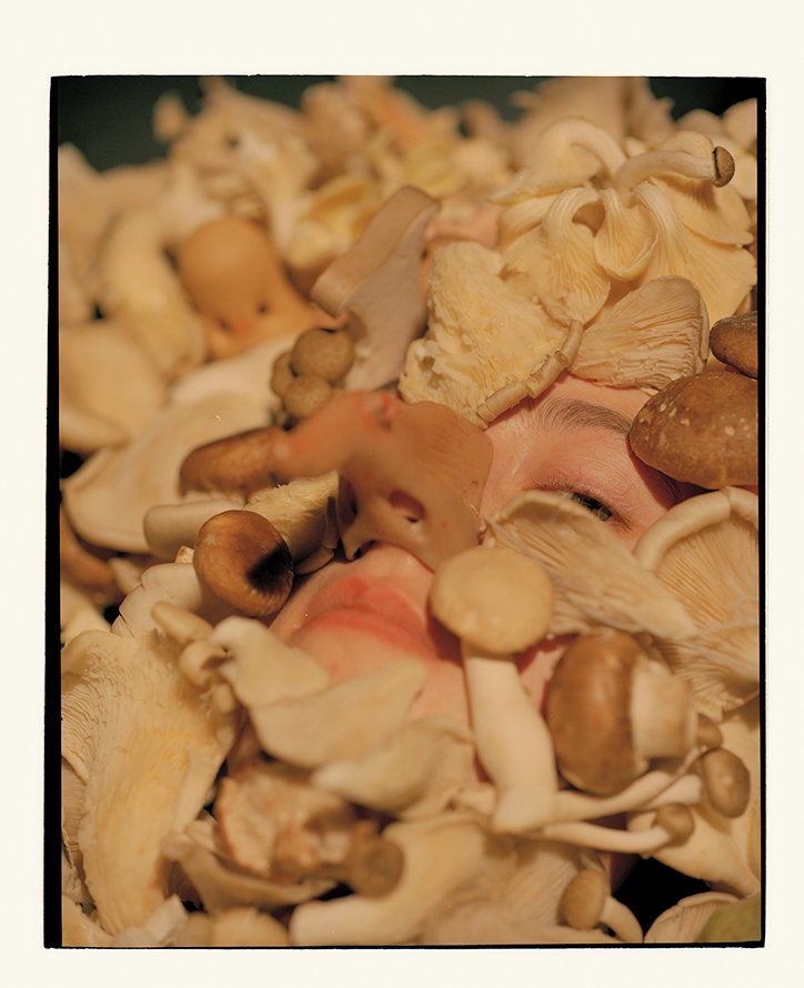 A close-up of a person’s face almost entirely covered in various types of pale mushrooms. Only one eye and part of the lips are visible through the fungi.