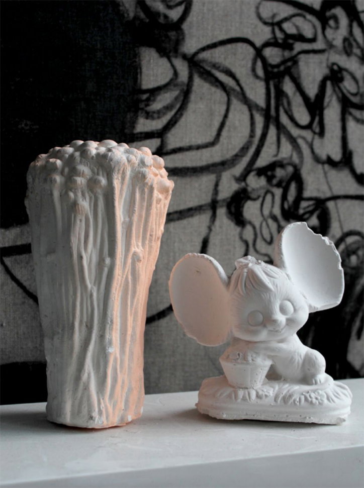 Two white ceramic sculptures sit on a plain white ledge. One is a tall, textured cylinder resembling a bundle of stalks, and the other is a small figure with large, rounded ears.