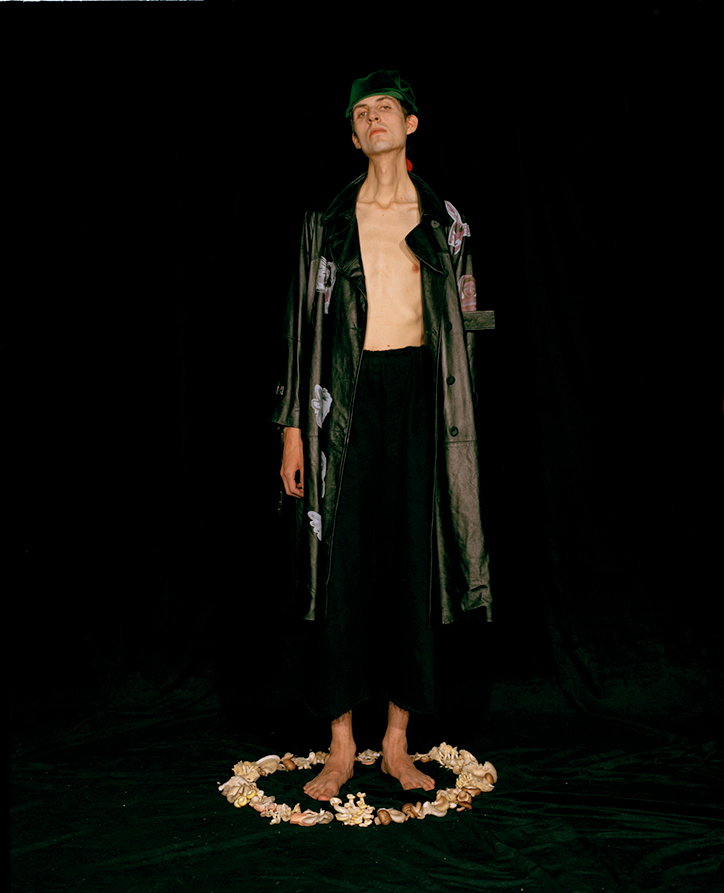 A person stands against a black backdrop, wearing a long dark coat over bare skin and a green cap. They stand barefoot inside a circle of seashells on a dark floor.