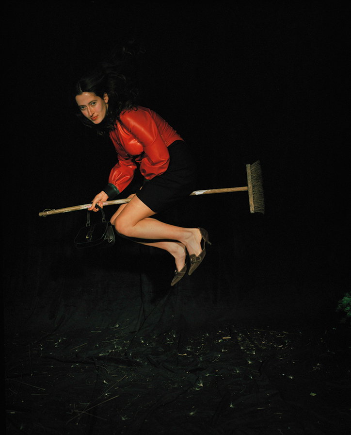 A person in a red top and black skirt leaps through the air against a black background. They hold a broom horizontally and carry a small black handbag.