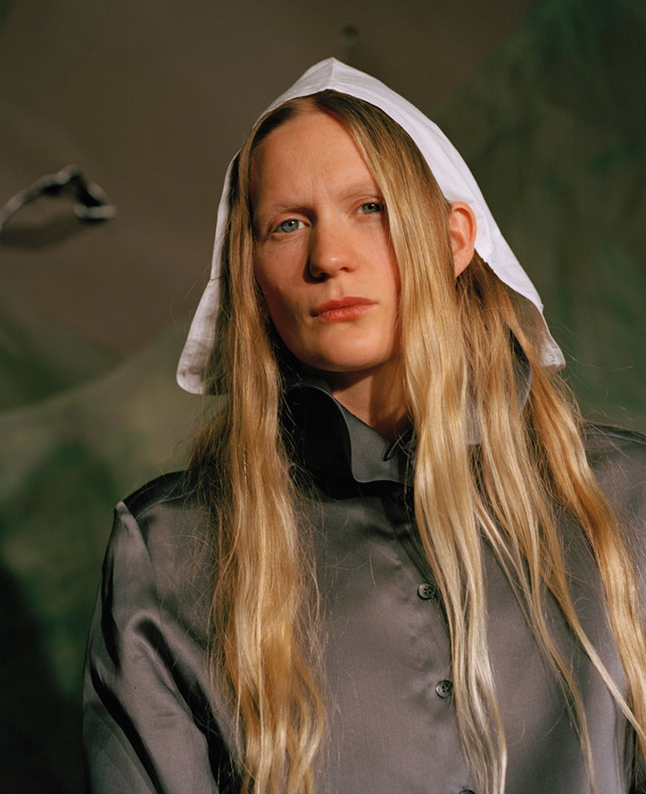 A person with long blonde hair looks directly at the camera. They wear a grey button-up shirt and a white, pointed head covering.