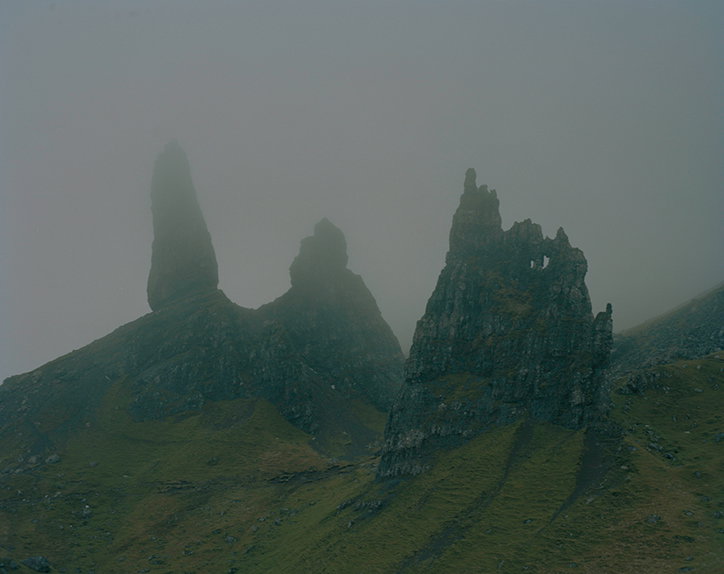 Three jagged rock pinnacles rise from a grassy hillside, partially obscured by a thick, grey mist. The landscape is muted and atmospheric, with the peaks fading into the fog.