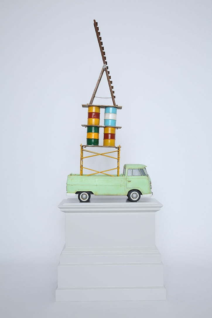 A sculpture of a light green vintage truck on a white plinth. A yellow scaffold rises from the truck bed, supporting four colourful drums and a tall wooden ladder.
