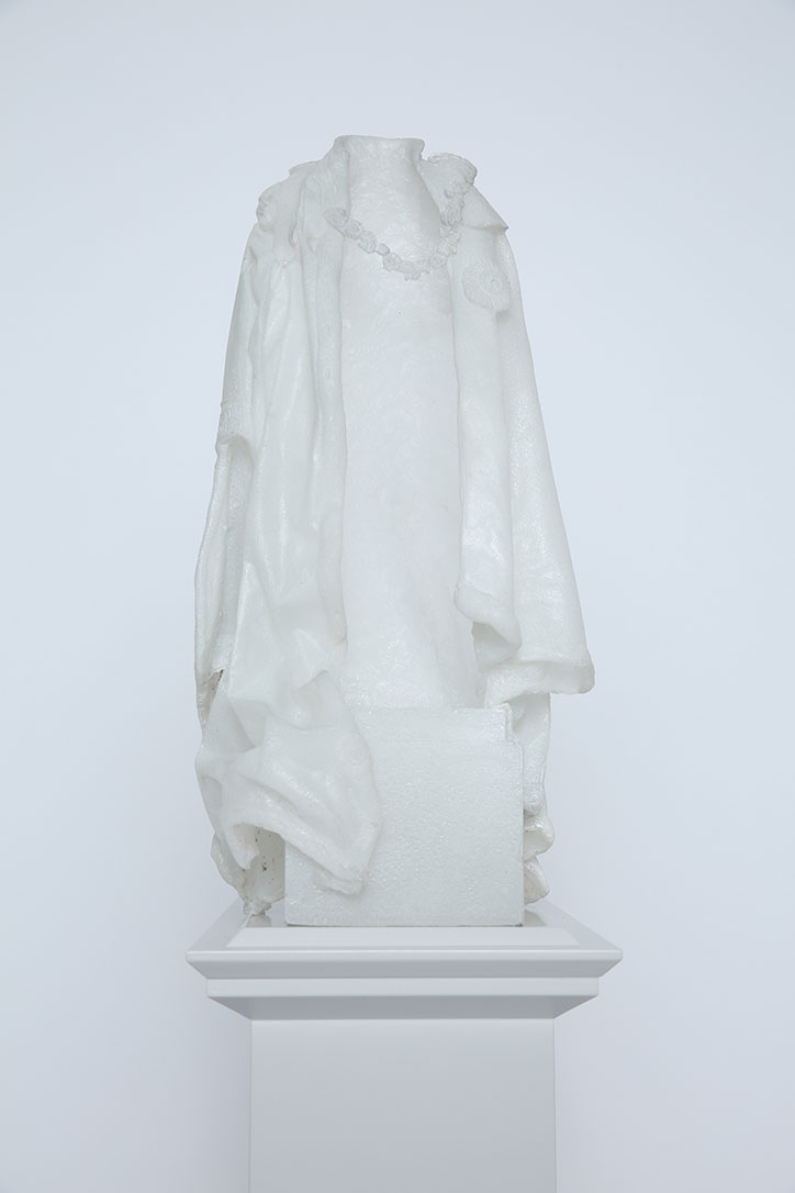 A white sculpture of a coat and a necklace, shaped as if worn by an invisible person. The piece stands on a white plinth against a plain white background.