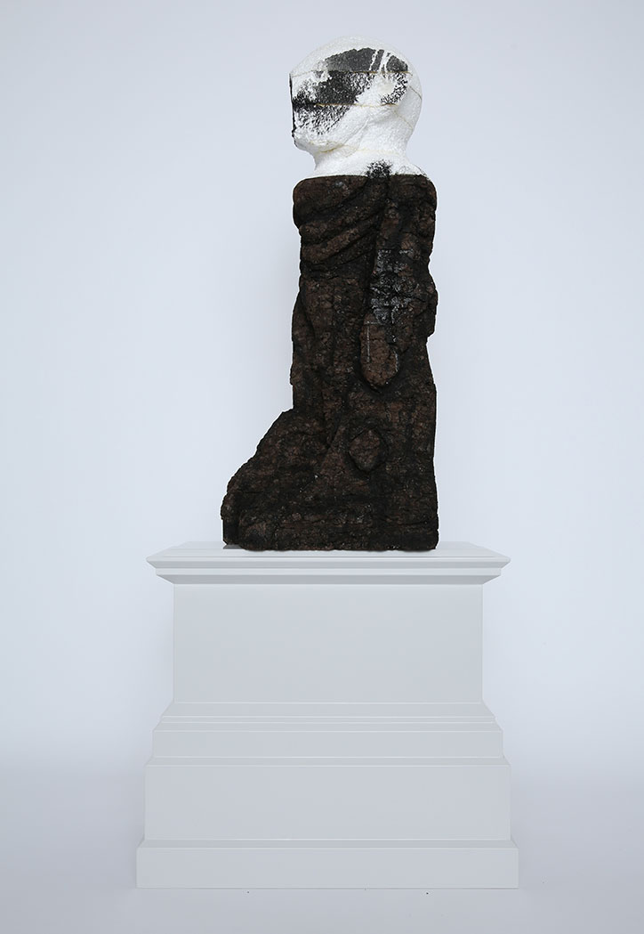 A sculpture of a standing figure on a white plinth. The body is a dark, textured mass, topped with a white head featuring a black painted band across the eyes.