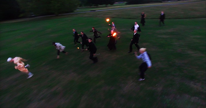 A group of people in formal black attire run across a dark green lawn. One person in the foreground wears a light-coloured outfit and leans back as they sprint.