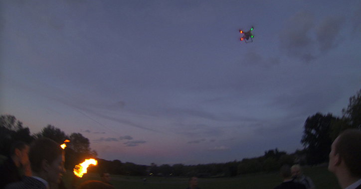 A drone with glowing red and green lights flies in a dark purple twilight sky. Below, a person performs with a flaming torch in a grassy field.