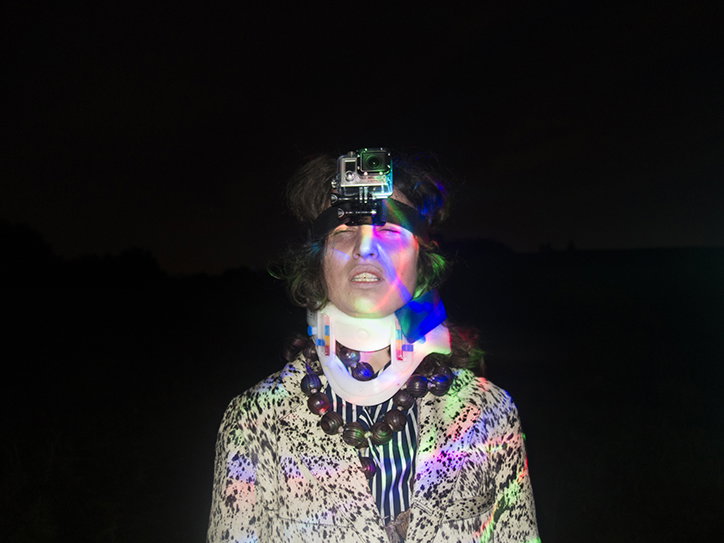 A person stands against a black background, wearing a small camera strapped to their forehead. Prismatic, rainbow-coloured light is projected across their face and a patterned jacket.