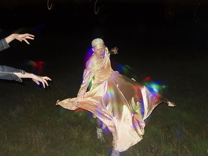 A person in a flowing gold gown and headpiece poses in a dark field at night. Prismatic light leaks in red, blue and green streak across the scene, while two hands reach in from the left.