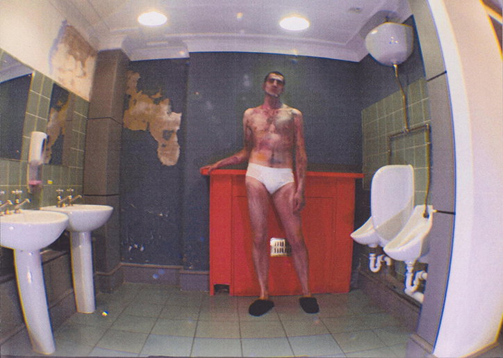 A person wearing white briefs stands in a dilapidated bathroom, leaning against a red partition. The walls are tiled in green and grey, with patches of plaster peeling away.