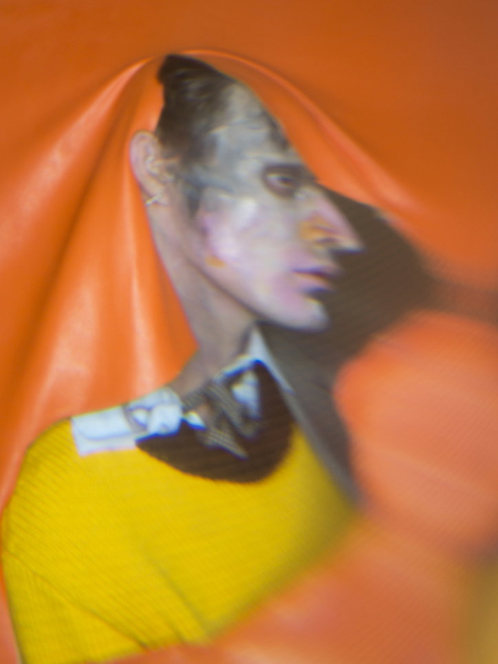 A person in a yellow top and white collar is seen through a fold in a bright orange fabric. Their face is viewed in profile, partially obscured by the draped material.