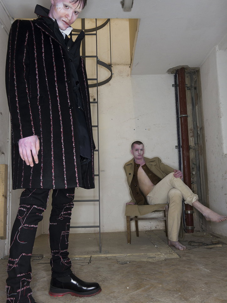 Two people with pale, stylised makeup pose in a sparse, industrial room. One person stands in the foreground wearing a long, black pinstriped coat and matching trousers, while another sits behind them in a tan jacket and trousers.