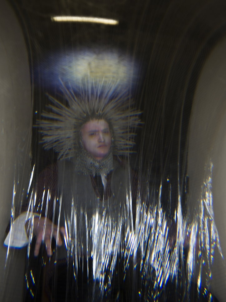 A person wearing a spiked, halo-like headpiece and a high collar looks directly at the camera. They are seen through a distorted, translucent plastic sheet that creates blurred vertical streaks across the frame.