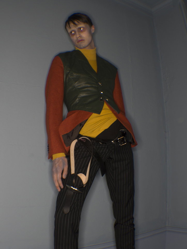 A person stands against a plain grey wall, wearing a yellow turtleneck under a dark green leather waistcoat and an orange jacket. They wear pinstriped trousers with a wide belt and a leather strap hanging from their hip.
