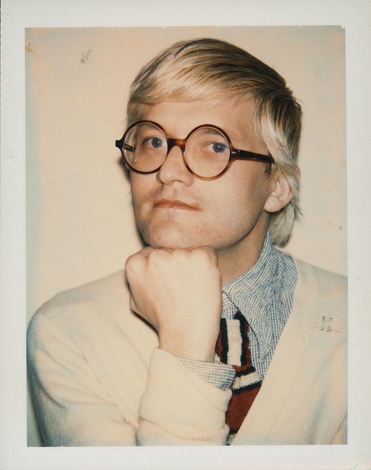 A person with blonde hair and round, tortoiseshell glasses looks directly at the camera. They rest their chin on a closed fist, wearing a light-coloured cardigan over a blue checked shirt and a striped tie.