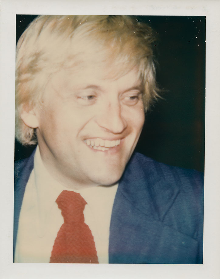 A person with blonde hair smiles, looking off-camera. They wear a blue blazer, a white shirt and a red tie.