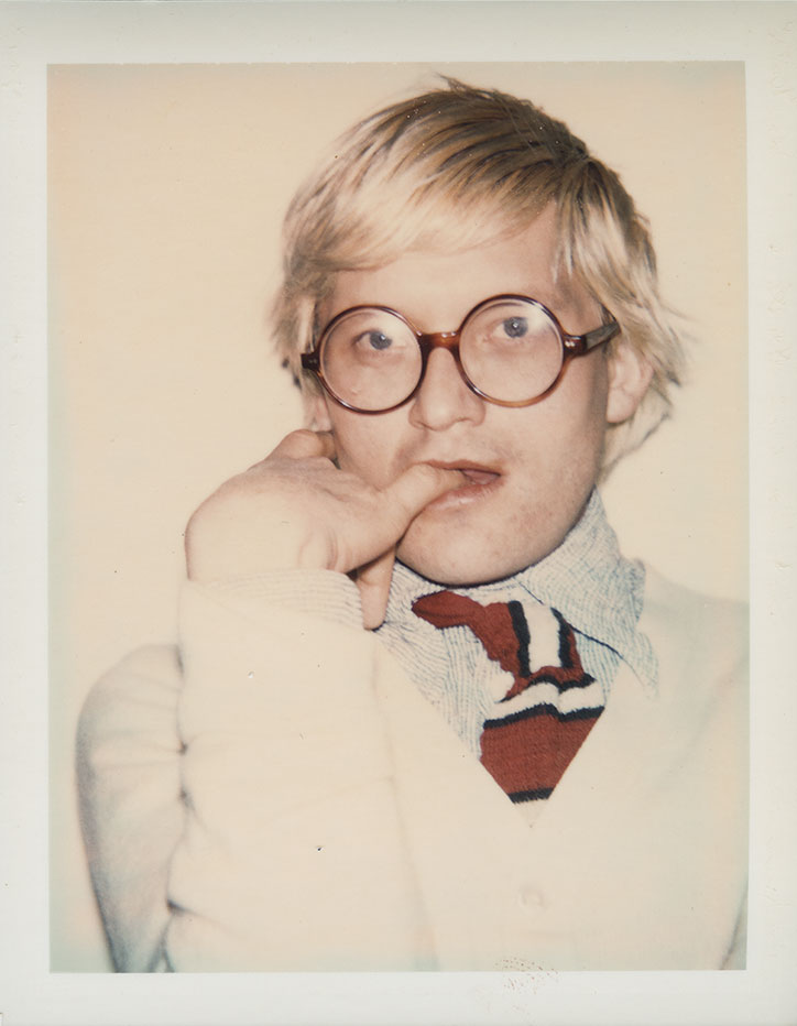 A person with blonde hair and round, dark-rimmed glasses looks at the camera. They wear a white jacket over a patterned shirt and a red and navy striped tie.