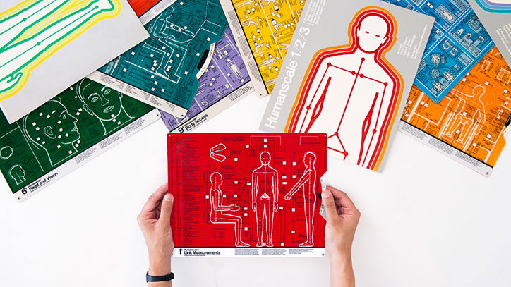Two hands hold a red technical diagram showing human figures in various poses. Around it, several other similar diagrams in green, blue, yellow and orange are scattered across a white surface.