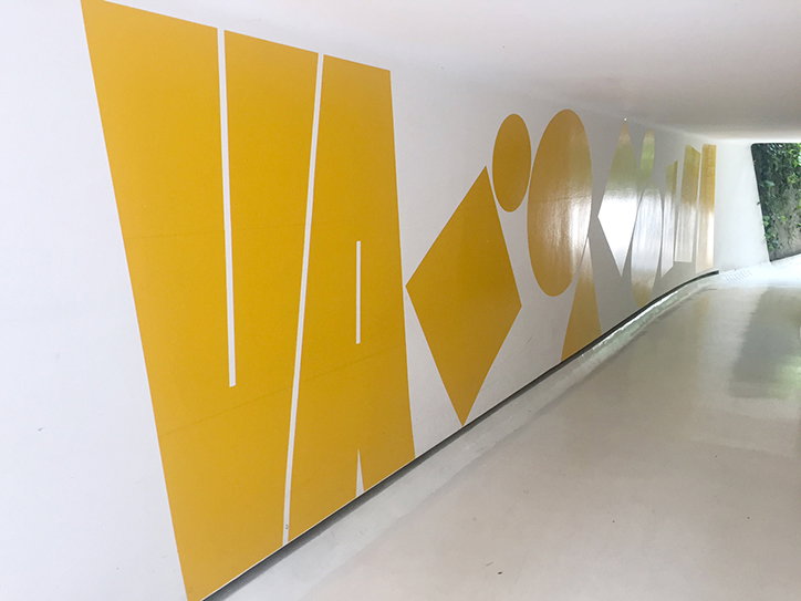 Large, yellow geometric shapes and abstract lettering are painted along a white wall in a bright corridor. The bold graphics create a rhythmic pattern that recedes into the distance.