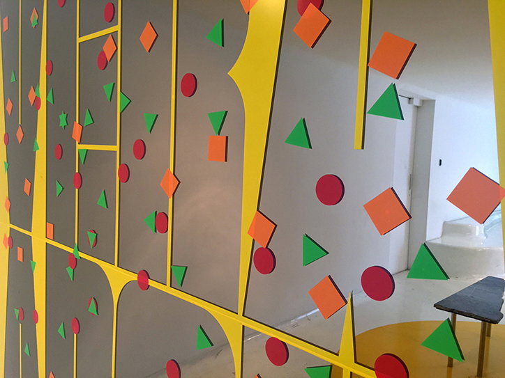 An installation of grey panels divided by thick yellow lines. Red circles, orange squares and green triangles are scattered across the surfaces.
