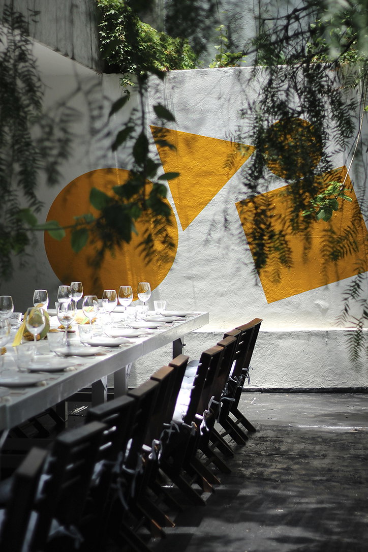 A long white dining table is set for a meal in an outdoor courtyard. Behind it, a white wall is decorated with large yellow geometric shapes.