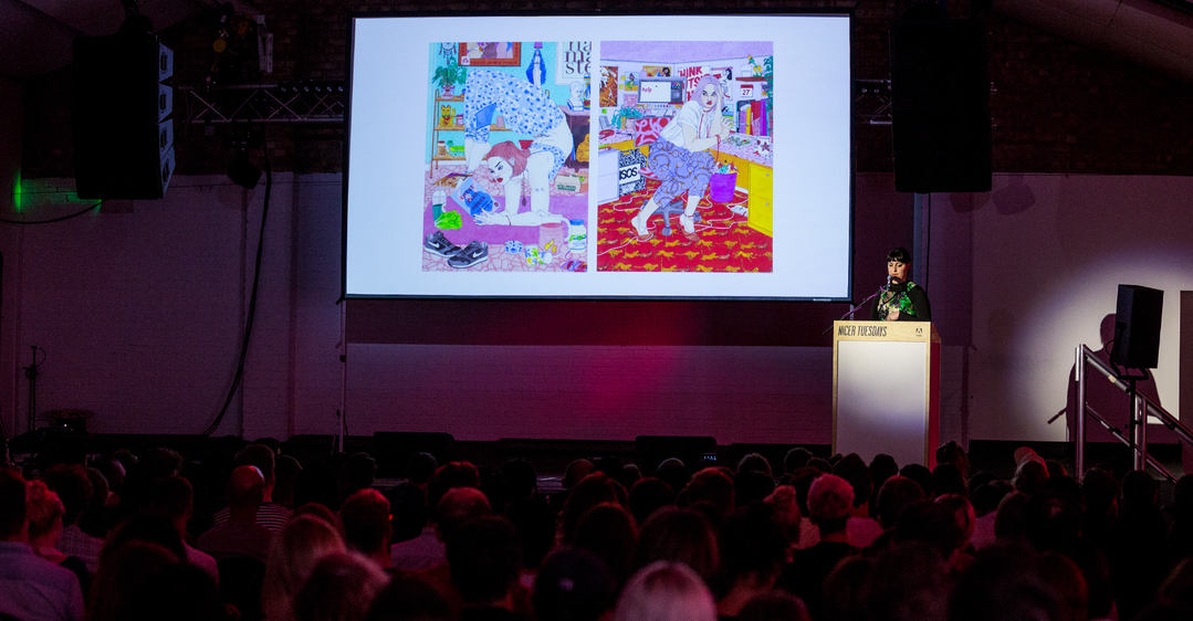 A person stands at a white lectern, speaking to a large audience in a darkened room. Behind them, a projection screen displays two colourful illustrations of figures in domestic settings.