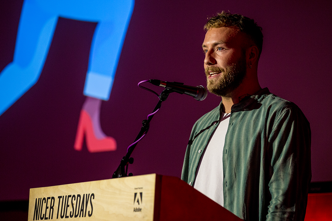 A person with a beard speaks into a microphone at a podium. The podium features the text “Nicer Tuesdays” and an Adobe logo.
