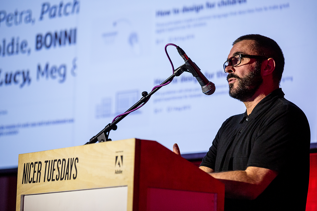 A person with glasses and a beard speaks into a microphone at a podium. The podium is branded with “Nicer Tuesdays” and the Adobe logo.