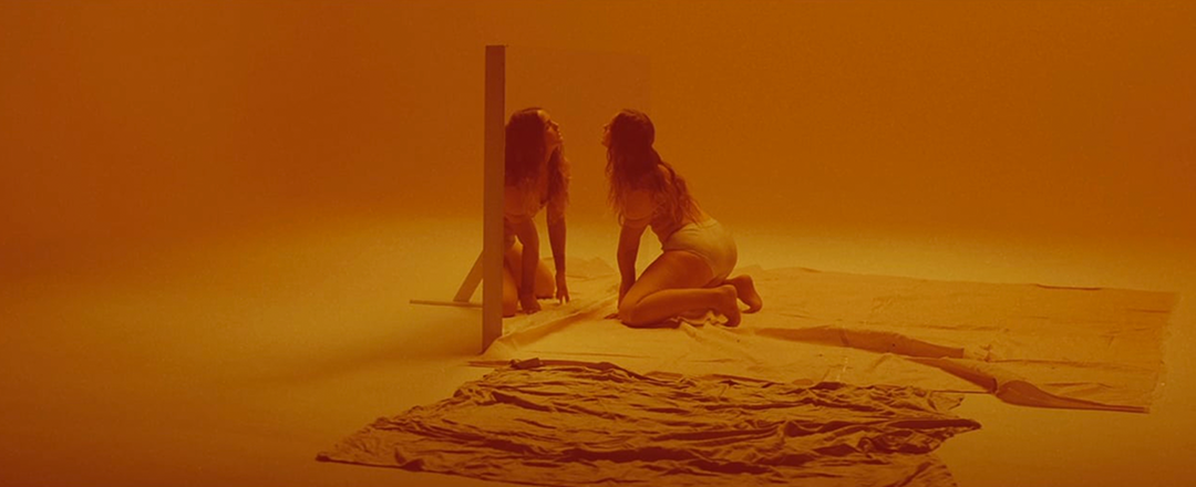 A person in white underwear kneels on a pale sheet, facing a large mirror. Their reflection is visible in the glass against a saturated orange background.