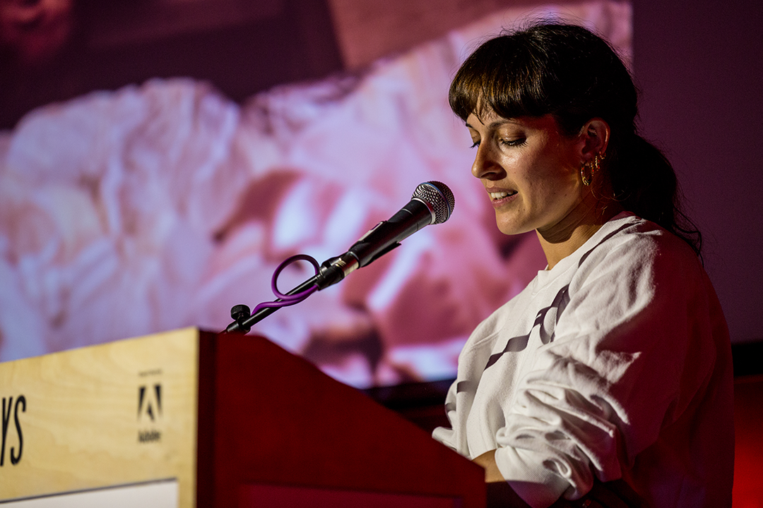 A person stands at a wooden lectern, looking down and speaking into a microphone. Behind them, a large screen displays a blurred, abstract image in shades of pink and white.