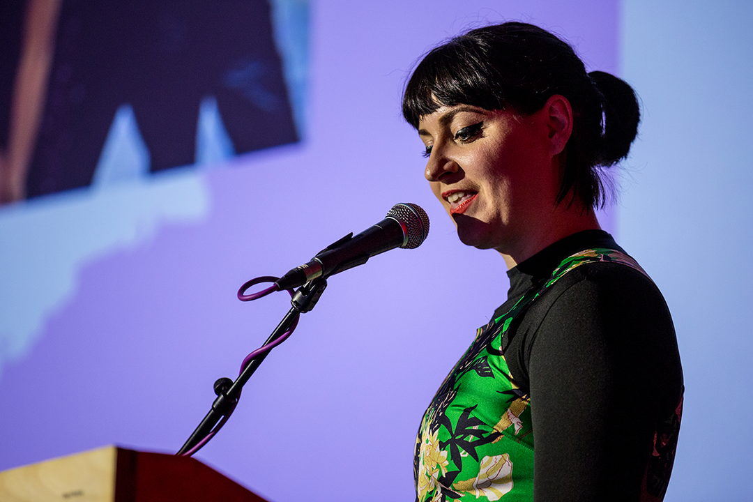 A person with dark hair and a fringe speaks into a microphone at a podium. They wear a black long-sleeved top under a green and yellow floral vest.