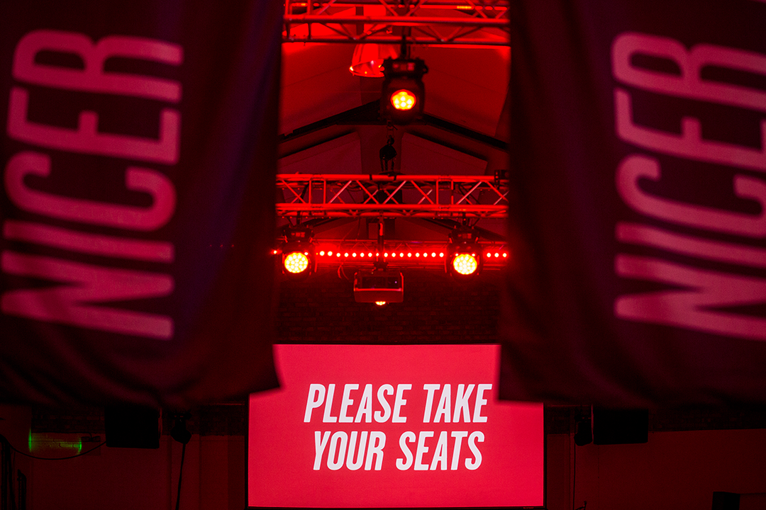 Two red banners with the word “NICER” frame a view of a stage. A bright red screen in the centre reads “PLEASE TAKE YOUR SEATS”.