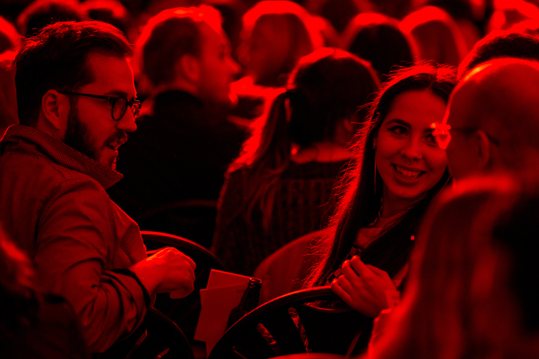 A crowd of people sit in a darkened room, bathed in deep red light. In the foreground, a person with glasses looks on from the left while another person smiles towards someone out of frame.