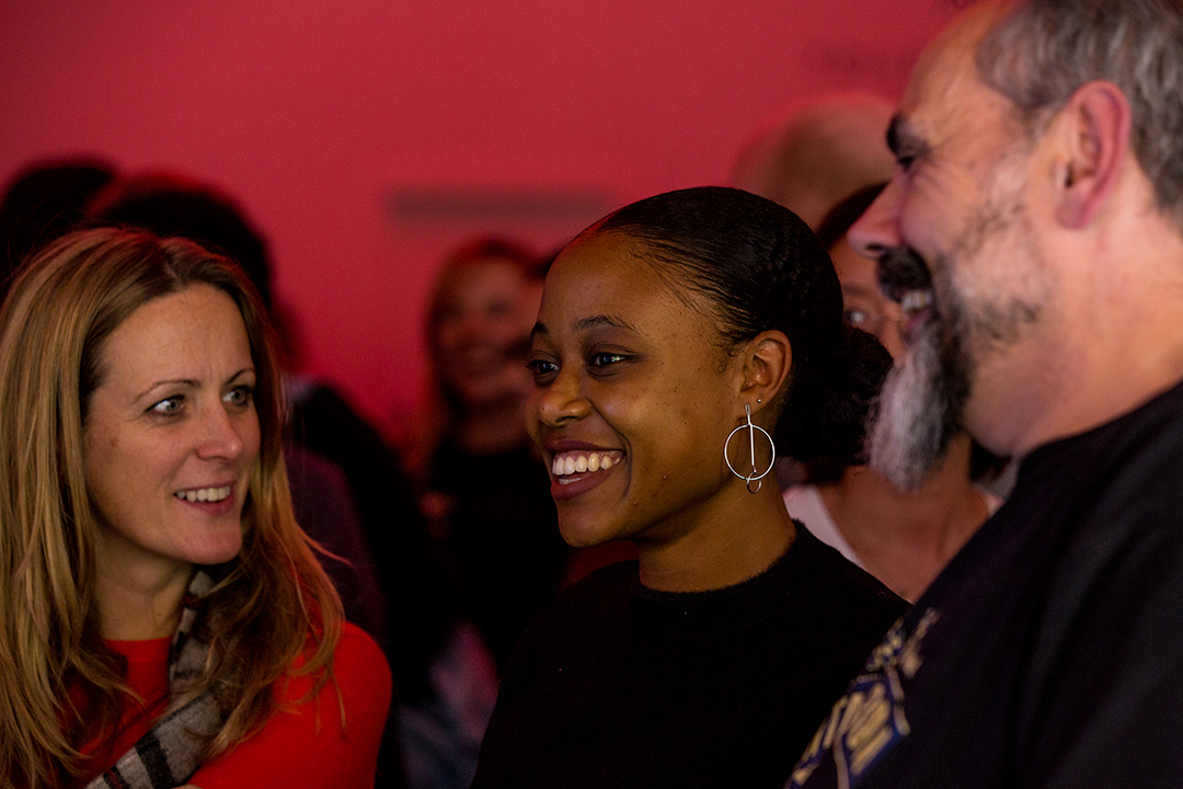 Three people stand together in a room with red lighting, engaged in conversation. A person in a black top smiles broadly while looking towards a person with a grey beard.