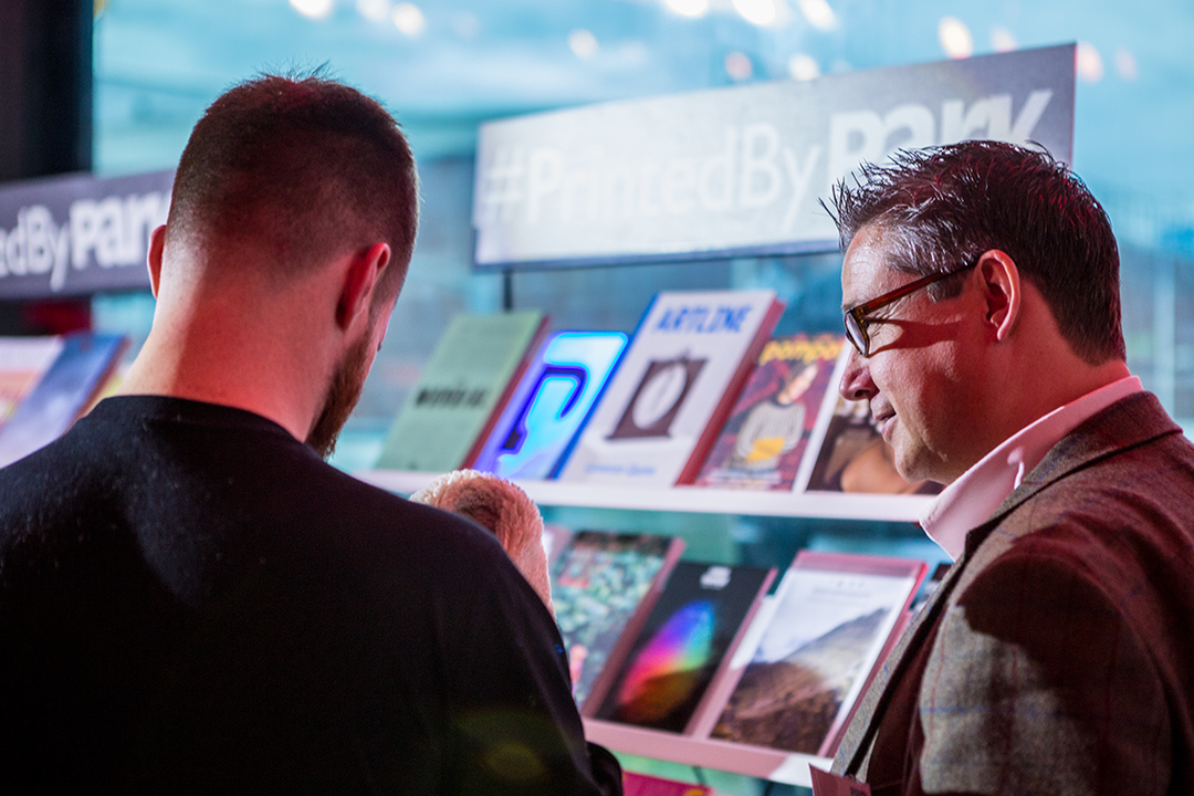Two people stand in a gallery, looking at a display of printed booklets and zines. A sign above the display reads “#PrintedByPark”.