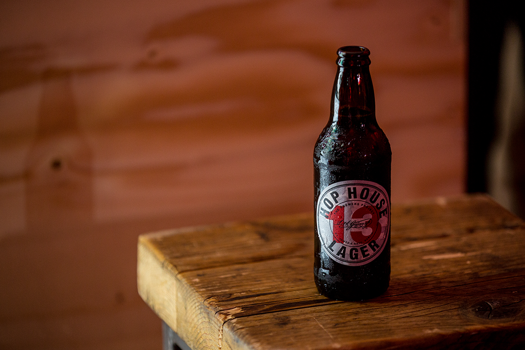 A condensation-covered brown glass beer bottle stands on a rustic wooden table. The label features a red circle with the number “13” and the words “Hop House Lager”.