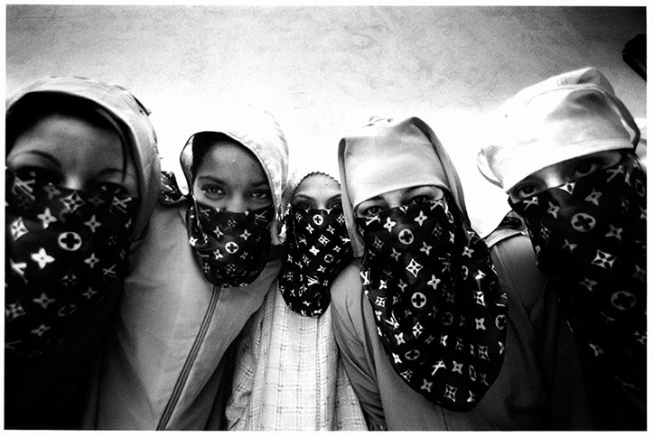 Five people lean in close to the camera, their faces partially covered by scarves featuring the Louis Vuitton monogram. They wear hoods or head coverings, with only their eyes visible.